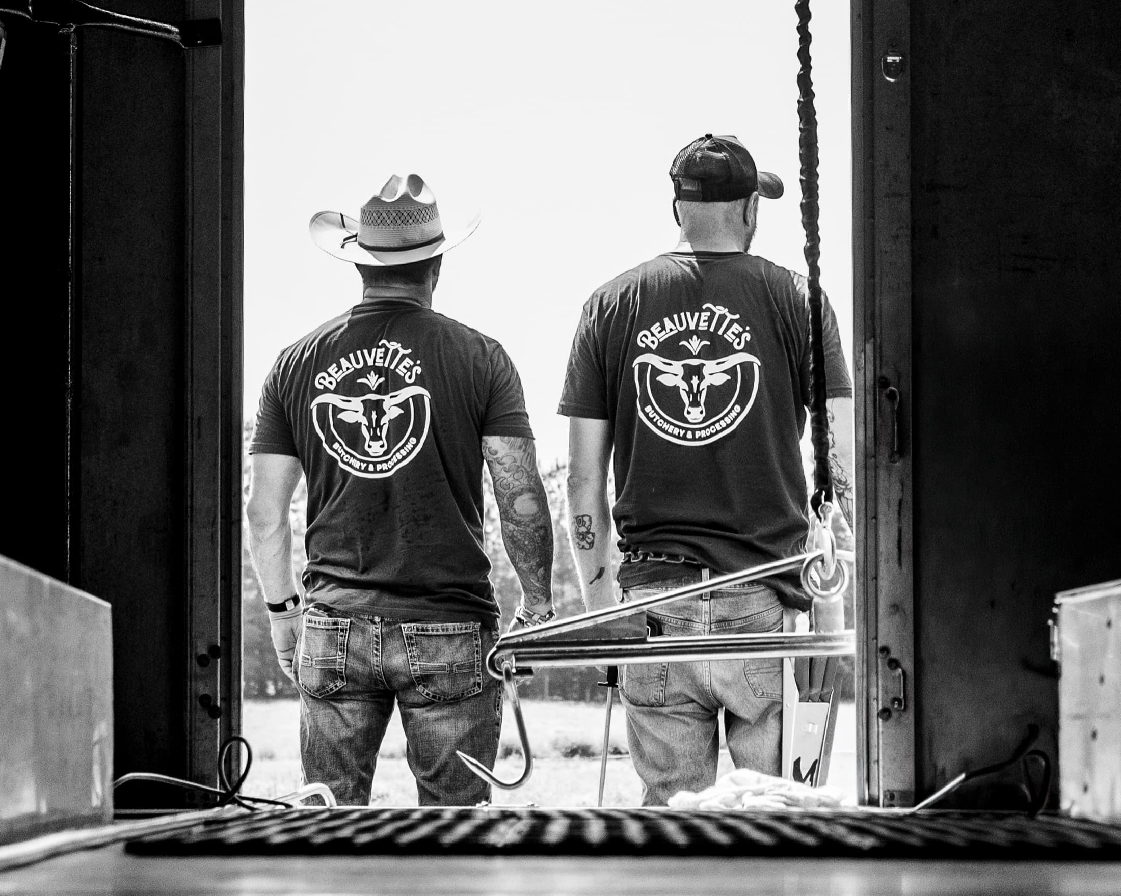Two Beauvette's butchers in branded shirts at the farm, seen from behind in a barn doorway